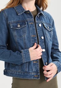 Veste en denim bleu au design classique, dotée de fournitures en métal argenté, de deux poches poitrine et d'une texture légèrement vieillie.