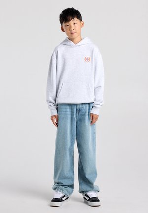 Jongen staat en draagt een lichtgrijze hoodie met voorzak, losse lichtblauwe spijkerbroek en zwarte en witte sneakers tegen een effen achtergrond.