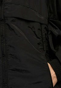 Giacca nera con zip realizzata in tessuto lucido e testurizzato con un design a strati, caratterizzata da un logo e polsini elastici. Dettagli minimalisti e tasche.