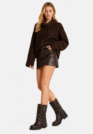 Donna che indossa un maglione oversize marrone scuro, una minigonna in pelle marrone scuro e stivali in pelle marrone scuro fino a metà polpaccio con fibbie.