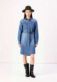 Denim srajčna obleka z dolgimi rokavi, sprednjimi gumbi in pasom. Ima svetlo modro pranje in podolgovato yoke podrobnost. Nosijo se z črnimi škornji do kolen.