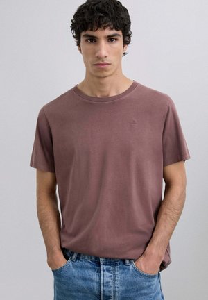 Camiseta corta de manga corta color marrón, hecha de algodón suave, con un escote redondo y un sutil detalle del logo en el pecho. Combinada con unos jeans azul claro.