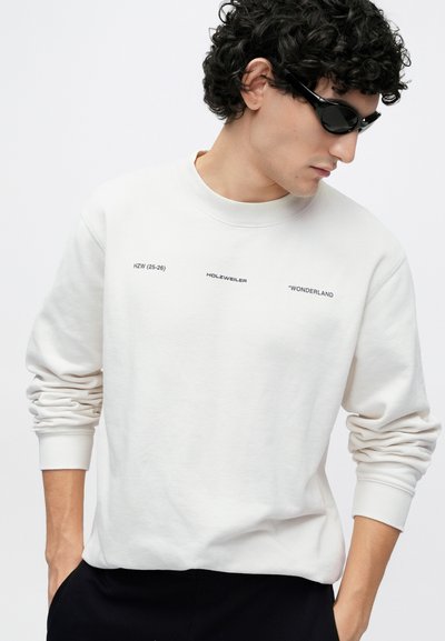 Svetlobéžový pulóver s okrúhlym výstrihom a dlhými rukávmi. Na prednej strane je čierny potlačený nápis: "HZW (25-26) HOLZWEILER 'WONDERLAND."
