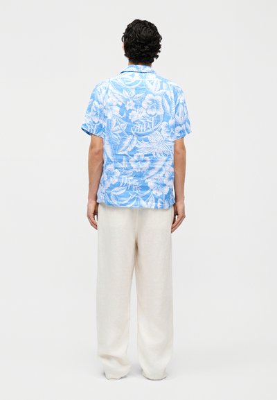 Egy férfi áll háttal, kék-fehér virágmintás rövid ujjú ingben és laza fehér nadrágban, egyszínű háttér előtt.