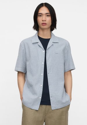 Person trägt ein kurzärmeliges, hellblau-weiß gestreiftes Hemd über einem schwarzen T-Shirt und beige Hosen, Hände in den Taschen.