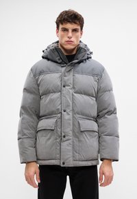 Chaqueta gris acolchada con capucha, que presenta un cierre de botones, dos bolsillos frontales y un panel superior en contraste. El material parece suave y con textura.