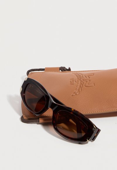 Γυαλιά ηλίου γατίσιου ματιού με κοχύλι χελώνης και σκούρους φακούς, τοποθετημένα πάνω σε δερμάτινη θήκη σε ταμπά χρώμα, με εγχάρακτο φύλλο δάφνης και το λογότυπο "MCM".