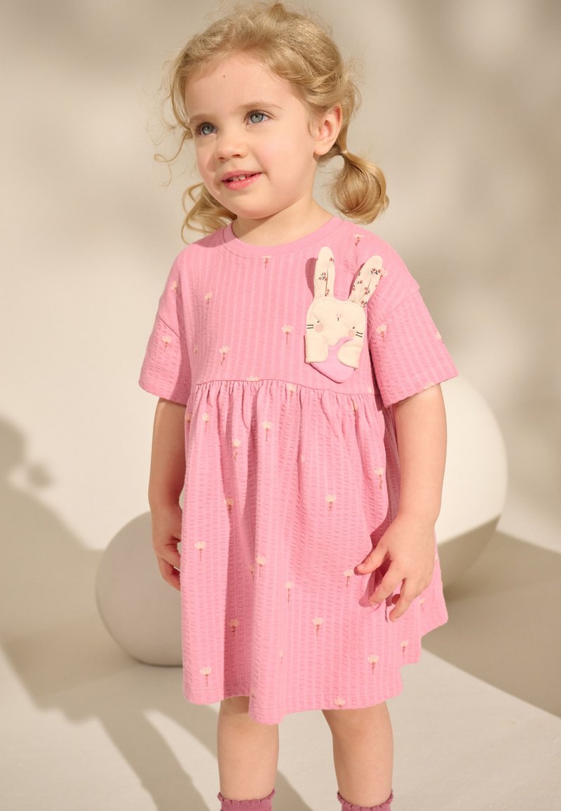 Peutermeisje met blond haar in vlechtjes, gekleed in een roze jurk met kleine bloemenprints en een pluche konijntje op de borst.