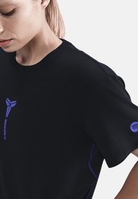 Kvinde med lyst hår iført sort FC Barcelona t-shirt med lilla logo på brystet og et lille lilla Spotify-logo på højre ærme.