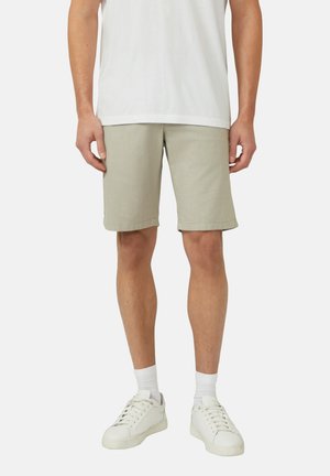 Mann trägt hellbeige gestreifte Shorts, weiße Sneakersocken, weiße Turnschuhe und ein weißes T-Shirt, steht vor einem schlichten Hintergrund.