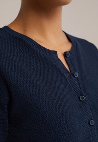 Tumesinine koerib kardigan, millel on tekstureeritud muster, ümara kaelusega ja viis nuppu esiosas. Sile ja pehme kangas.