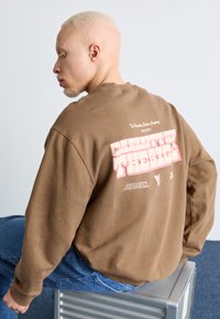 Sudadera marrón con un gran gráfico rosa en la parte posterior que incluye texto. La tela tiene una textura única, y la sudadera tiene un corte relajado y puños acanalados.