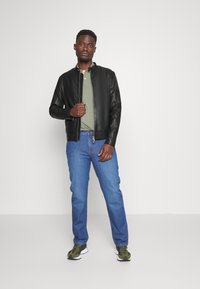 Zwarte leren bomberjack over een groene polo, gecombineerd met blauwe recht-toe-rechtaf jeans en groene camouflage sneakers. Eenvoudige, casual outfit.