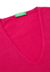 Maglione rosa acceso a scollo a V con etichetta verde "United Colors of Benetton" cucita al collo su uno sfondo bianco.