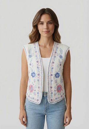 Femme portant un gilet blanc sans manches avec une broderie florale bleue et rose sur un haut blanc et un jean bleu clair, debout devant un fond uni.