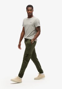Camiseta gris de manga corta combinada con pantalones cargo verde oliva que tienen bolsillos laterales, además de unas zapatillas bajas beige. Conjunto sencillo y casual.