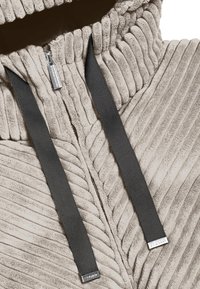 Beżowy, prążkowany polarowy hoodie z przednim zamkiem błyskawicznym i ciemnoszarymi sznurkami z akcentami marki. Miękka faktura i zorganizowany krój.