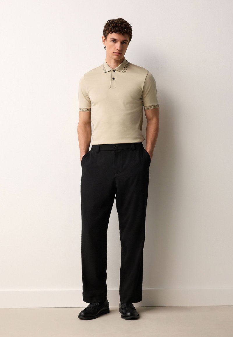 Beiges Poloshirt mit gemusterten Manschetten, kombiniert mit schwarzen Hosen. Das Modell steht aufrecht, die Hände in den Taschen, und trägt schwarze Schuhe.
