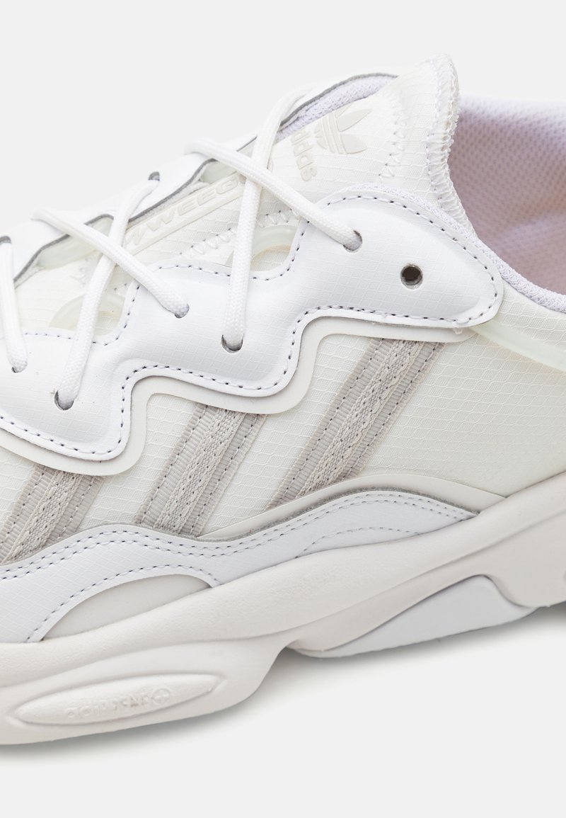 Białe sneakersy z teksturowanej siateczki, z trzema subtelnymi beżowymi paskami i zaokrągloną, rzeźbioną białą podeszwą. Posiadają płaskie białe sznurowadła i branding.