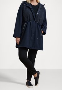 Navy quilted jacket with a drawstring waist, hood, and zippered pockets, paired with black leggings and dark sneakers. Smooth texture.