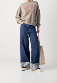 Sudadera gris de gran tamaño con gráfico rojo, combinada con vaqueros azul ancho con dobladillo deshilachado y zapatillas negras. Bolso tote beige.