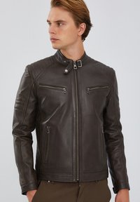 Giacca in pelle marrone con colletto alto, chiusura con zip e quattro tasche con zip. Presenta dettagli trapuntati sulle spalle. Tessuto liscio.
