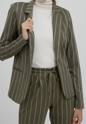 Femme portant une veste de costume à fines rayures vert olive et un pantalon assorti sur un col roulé blanc, tenant le revers de la veste avec la main gauche.