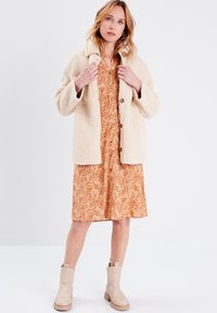 Cremefarbene Kunstschaffelljacke über einem orangefarbenen Blumenkleid mit Knöpfen. Beigefarbene Ankle Boots mit strukturiertem Finish und robuster Sohle.