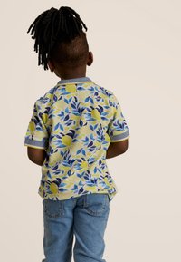 Korte mouwen polo in geel met een blauw blad- en citroenpatroon, met een blauwe kraag en manchetten. Draagt men met lichtblauwe jeans.