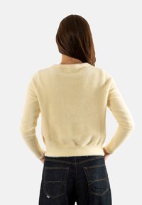 Suéter cremoso y esponjoso de mangas largas y cuello redondeado, combinado con jeans oscuros. El suéter tiene una cintura ceñida y una textura suave.