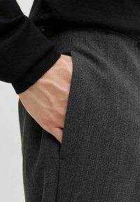 Pantalones grises a medida hechos de tela suave, con un bolsillo lateral y una mano inserta, acompañados de un suéter de punto negro.