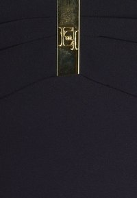 Ingrandimento di un tessuto nero con cuciture curve e un emblema rettangolare verticale in metallo dorato, impreziosito dalle lettere "EH".