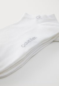 Fehér pamut zoknik egymásra rakva, bordás szegéllyel és szürke "Calvin Klein" logóval az egyik oldalon, sima textúrával, klasszikus crew hosszúságban.