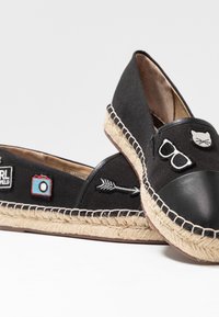 Espadrilles noires avec semelles en corde beige ornées de patchs brodés représentant un visage de chat, des lunettes de soleil, une flèche, un appareil photo et un patch logo.