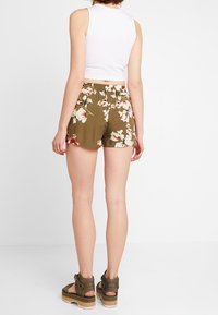 Shorts à fleurs vert olive en tissu léger avec taille élastique. Associés à un haut blanc sans manches et des sandales à plateforme.