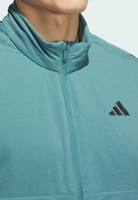 Veste de sport teal avec un col haut, fermeture éclair à l'avant et un détail de logo noir. Fabriquée à partir d'un tissu lisse et léger avec une texture subtile.