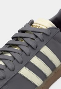 Γκρι αθλητικό παπούτσι με μπεζ λεπτομέρειες, με κλασικές τρεις ρίγες. Υλικό με υφή, κορδόνια σε αντίθεση και σόλα από καουτσούκ. Χρυσό λογότυπο στη γλώσσα.