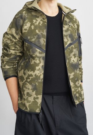 Man draagt een open groene en zwarte digitale camouflagejas over een zwart shirt en zwarte broek, handen in de zakken.