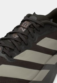 Σκούρο καφέ αθλητικό παπούτσι με ανοιχτό γκρι ρίγες, υφασμένο επάνω μέρος και εμφανές λογότυπο στη γλώσσα. Οι κορδόνια είναι σκούρα και επίπεδα.