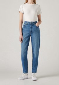 Jeans in denim blu a vita alta con gamba dritta, dotati di cinque tasche, passanti per cintura e chiusura con bottone classico. Abbinati a una maglietta bianca.