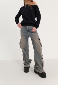 Sweat-shirt noir à épaules dénudées associé à un pantalon cargo ample bleu clair avec poches latérales, complété par des chaussures noires épaisses.