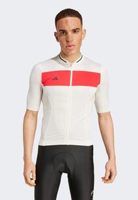 Jersey de ciclismo blanco con mangas cortas, que presenta una franja roja horizontal y acentos negros. Combinado con pantalones cortos de ciclismo negros.