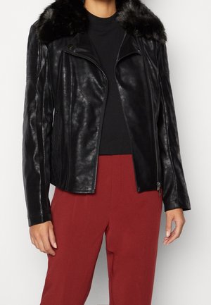 Veste en cuir noire avec col en fausse fourrure portée sur un haut noir et associée à un pantalon taille haute couleur rouille sur une personne debout.