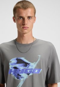 Gray cotton T-shirt featuring a blue graphic print with the word "HUGO." Short sleeves, crew neck, and a smooth texture. Silver chain necklace.
