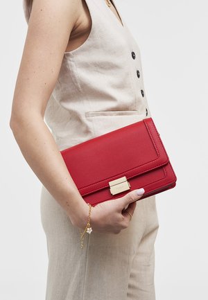 Kvinde i beige ærmeløs vest, der holder en rød læderclutch med en gylden lås og bærer et armbånd med gyldne charms.