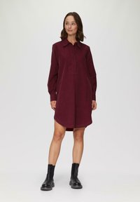 Langärmliges Hemdkleid in Burgunderrot, aus weichem Stoff gefertigt. Verfügt über eine Knopfleiste, einen spitzen Kragen und einen abgerundeten Saum. Wird mit schwarzen Stiefeletten getragen.