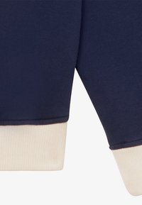 Tengerészkék pulóver krémszínű bordás mandzsettákkal, puha anyagból és laza fazonnal. A mandzsettákon kontrasztos színű részletek találhatók.