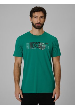 Mężczyzna z brodą, ubrany w zielony t-shirt z krótkim rękawem z grafiką o tematyce koszykarskiej i czarne spodnie, stojący na jednolitym szarym tle.