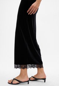 Fekete bársony hosszú ruha csipke díszítéssel az alján. A ruha egyenes szabású, és fekete pántos magassarkú szandállal van párosítva.