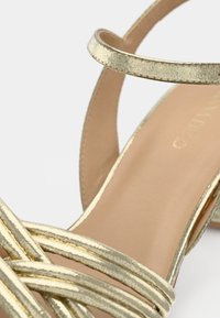 Χρυσό μεταλλικό πέδιλο με τρεις λεπτές μπροστινές λωρίδες και λουράκι στον αστράγαλο πάνω σε ταμπά εσωτερική σόλα με λευκό φόντο.